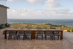 Ein langer Holztisch mit sechs Korbstühlen auf einer Steinterrasse mit Blick auf eine Küstenlandschaft, Hügel und das Meer.