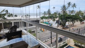 Un balcone con un tavolo e sedie in vetro, affacciato su una spiaggia e sullo skyline urbano, con palme e edifici.