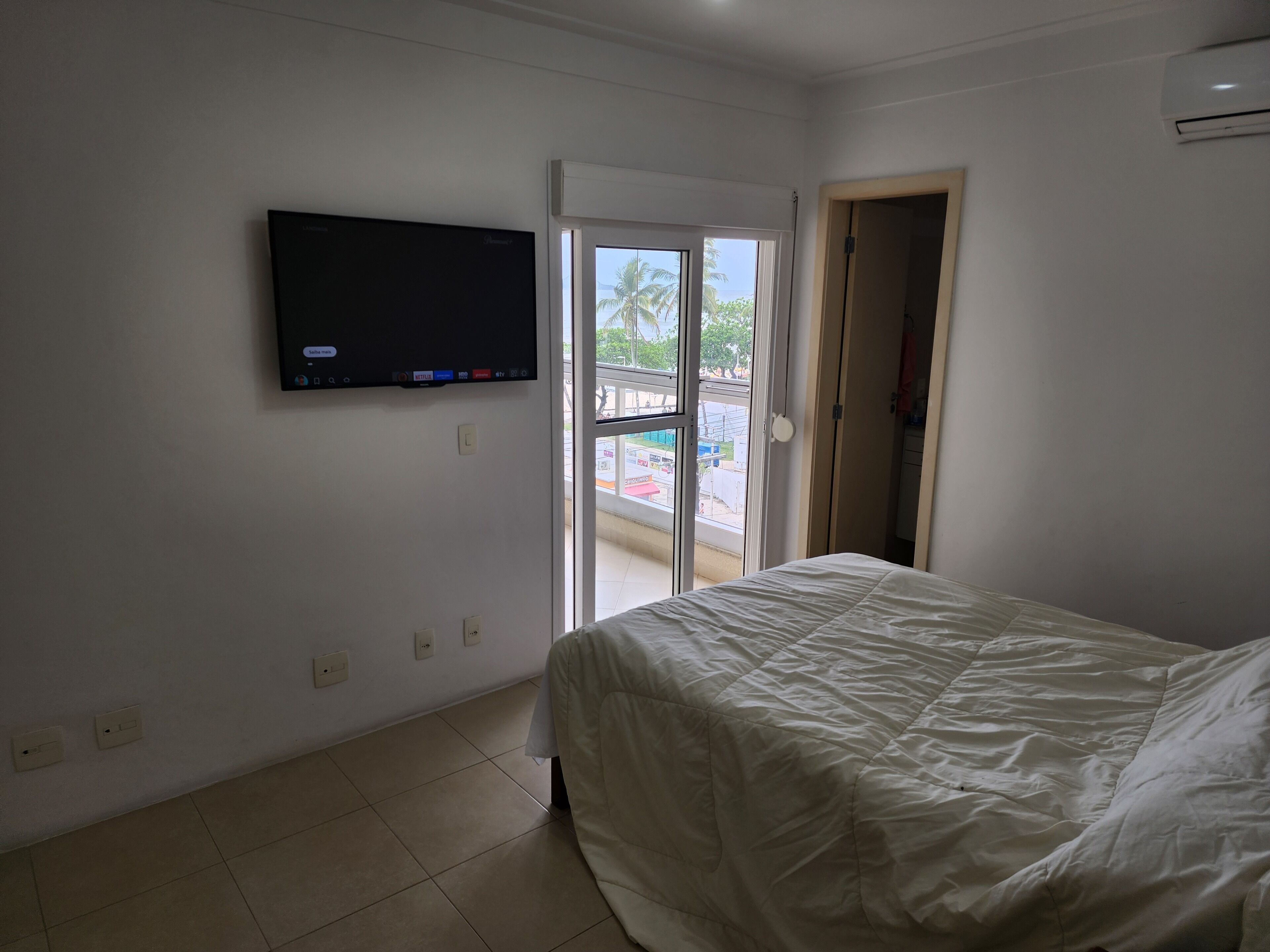 A bedroom with a bed, a TV mounted on the wall, and a sliding glass door leading to a balcony.