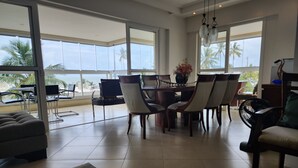 Un'ampia sala da pranzo con un tavolo e sedie di grandi dimensioni, ampie finestre che si affacciano su palme e sull'oceano, e un design moderno.