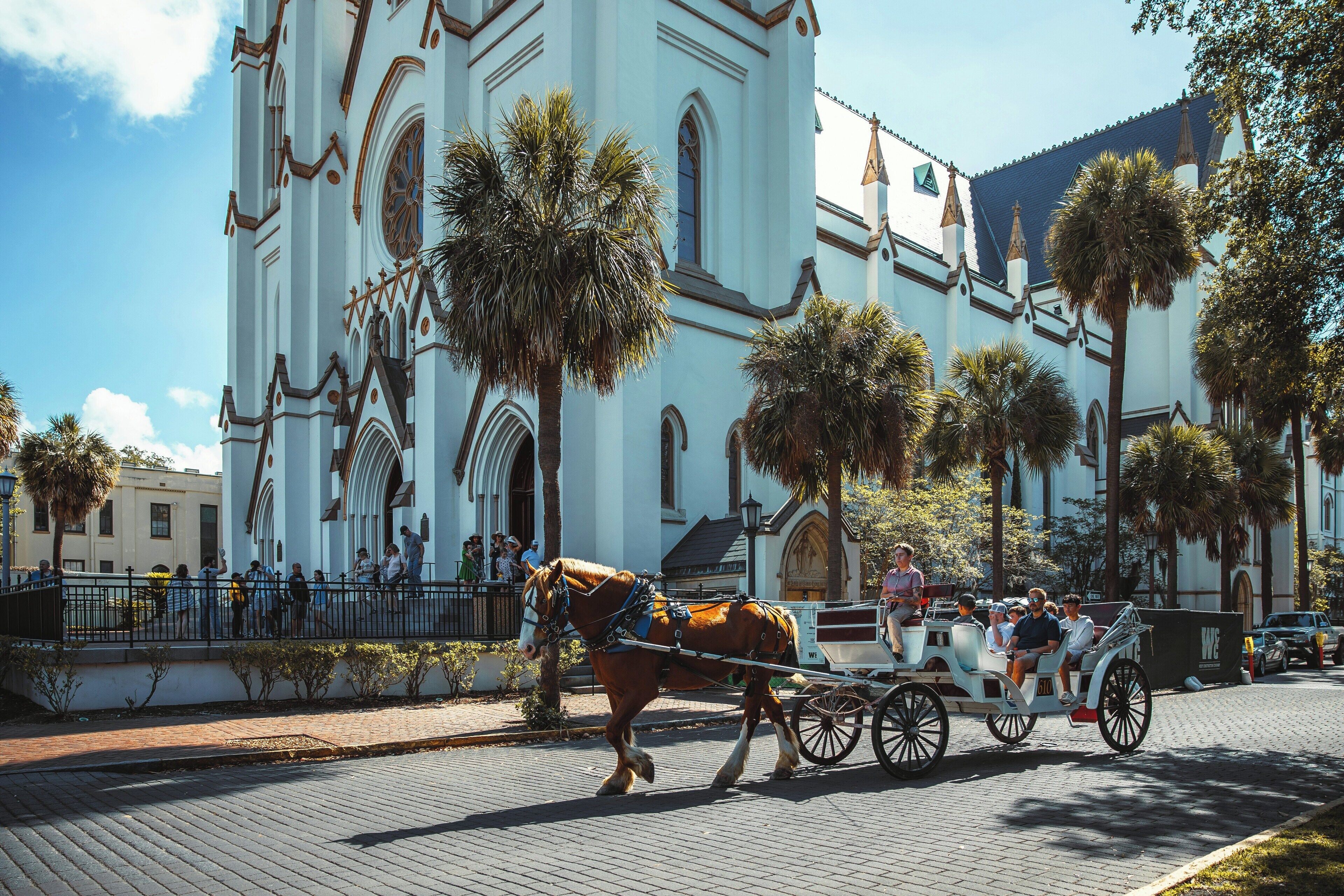 A horse-drawn carriage with passengers in front of a white church with arched windows and a bell tower.