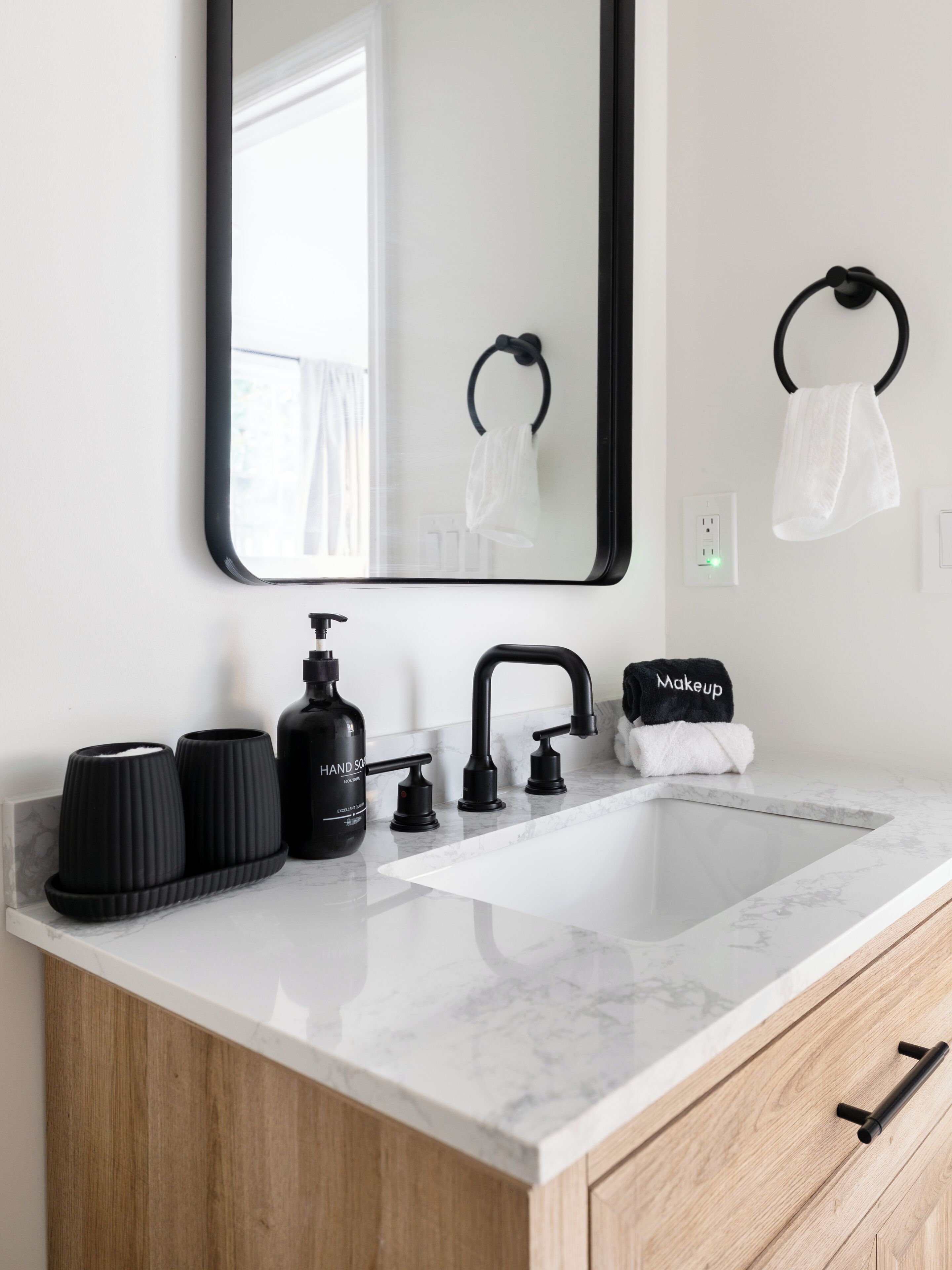 Un baño moderno con un lavamanos de mármol blanco, grifo negro y dispensador de jabón negro. Hay dos jaboneras negras y un tozalengua negro con una toalla blanca.