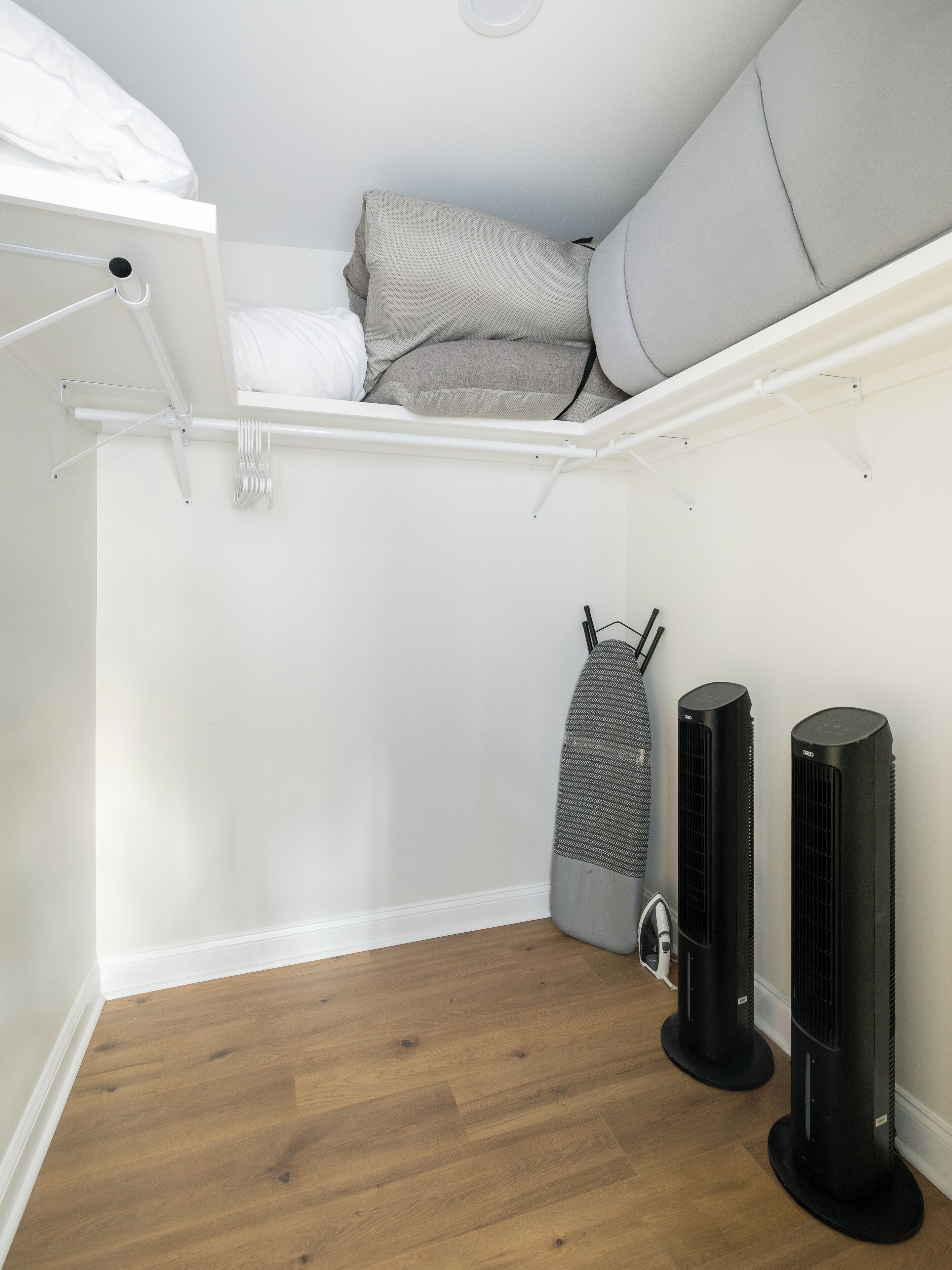 Ein kleines Zimmer mit Holzboden, einem Bett mit weißer Bettwäsche, einem grauen Bügelbrett und zwei schwarzen Ventilatoren.
