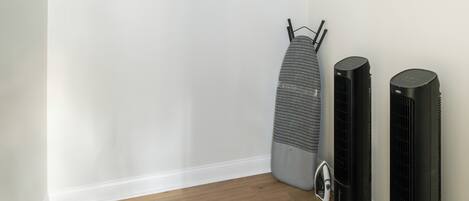 Ein kleines Zimmer mit Holzboden, einem Bett mit weißer Bettwäsche, einem grauen Bügelbrett und zwei schwarzen Ventilatoren.