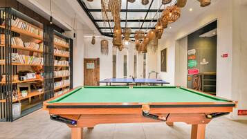 A billiards table with a green felt surface, surrounded by bookshelves and hanging wicker baskets.