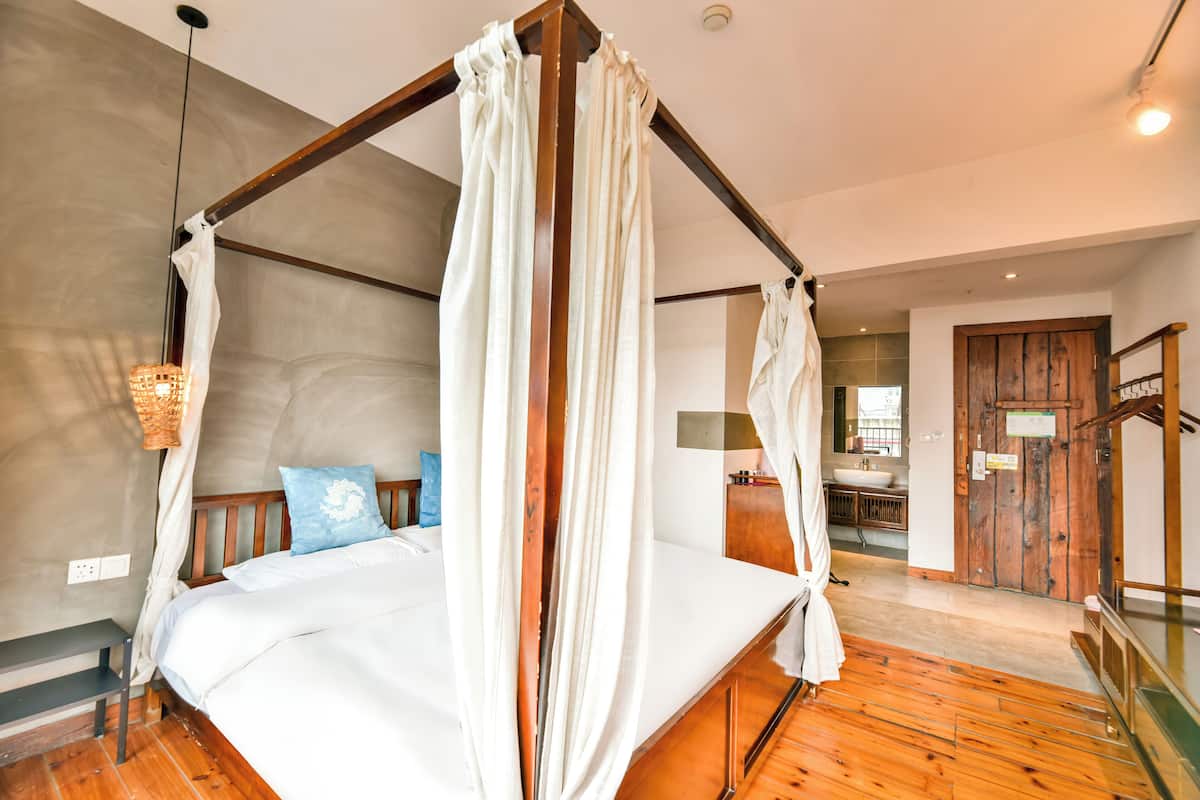 Uma cama de dossel com lençóis brancos e um travesseiro azul, um banco de madeira, um lustre pendurado e piso de madeira.