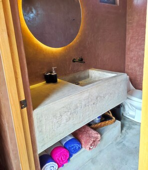 Ein Badezimmer mit einem großen, runden Spiegel, einem Steingefäß als Waschbecken und einem Regal mit Handtüchern.