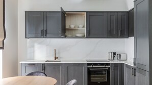 Uma cozinha moderna com armários cinza, bancada branca, forno embutido e área de jantar com mesa de madeira e duas cadeiras.