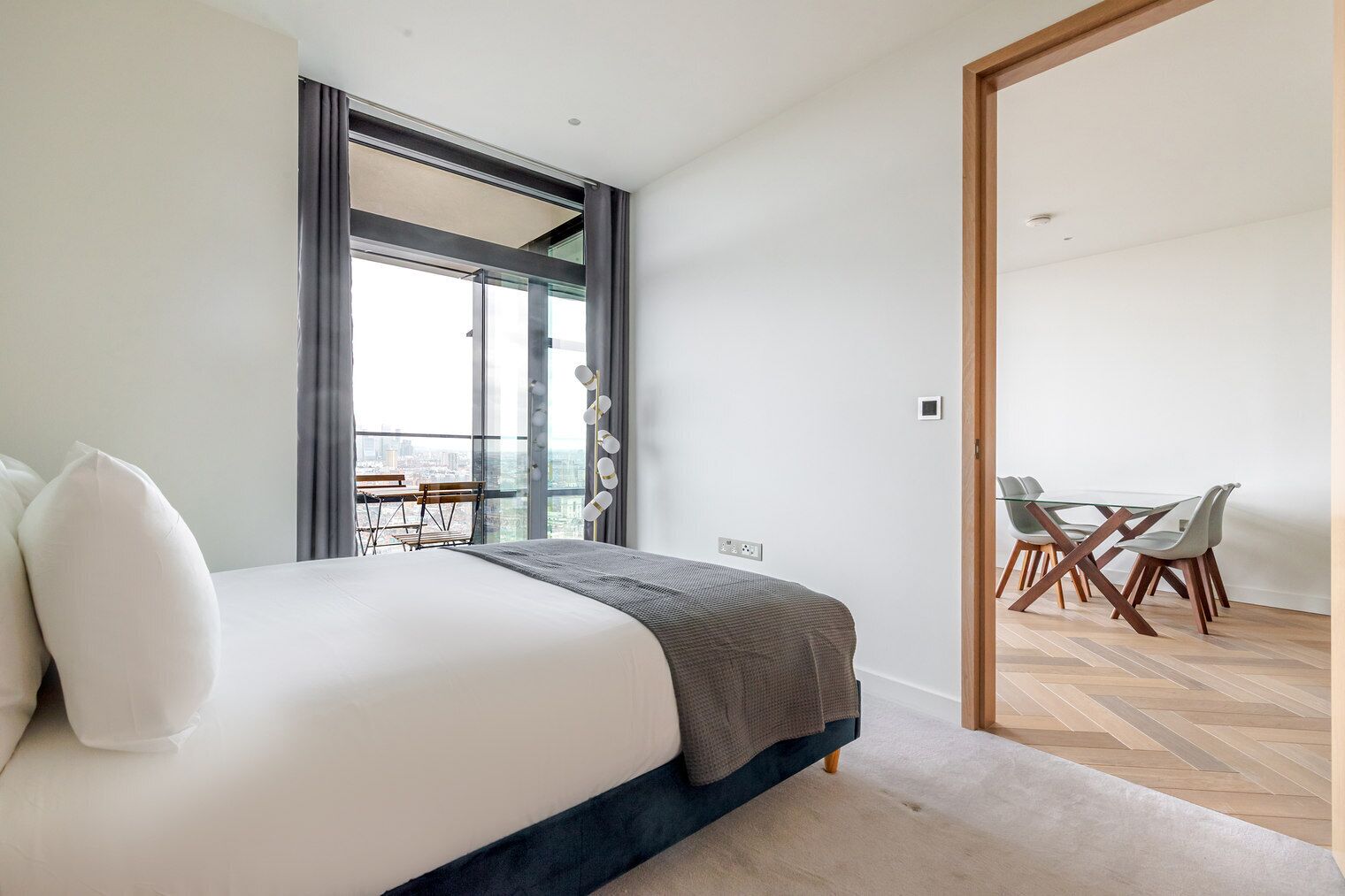 A modern bedroom with a large bed, a view of the city through a sliding door, and a dining area with a table and chairs.