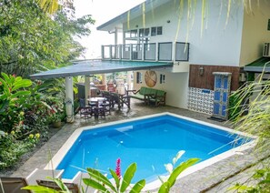 Ein Pool mit überdachtem Patio, umgeben von üppiger Bepflanzung und einem zweistöckigen Gebäude mit Balkon.