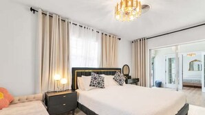 A modern bedroom with a large bed, a nightstand with a lamp, and a view of the living area through a glass door.