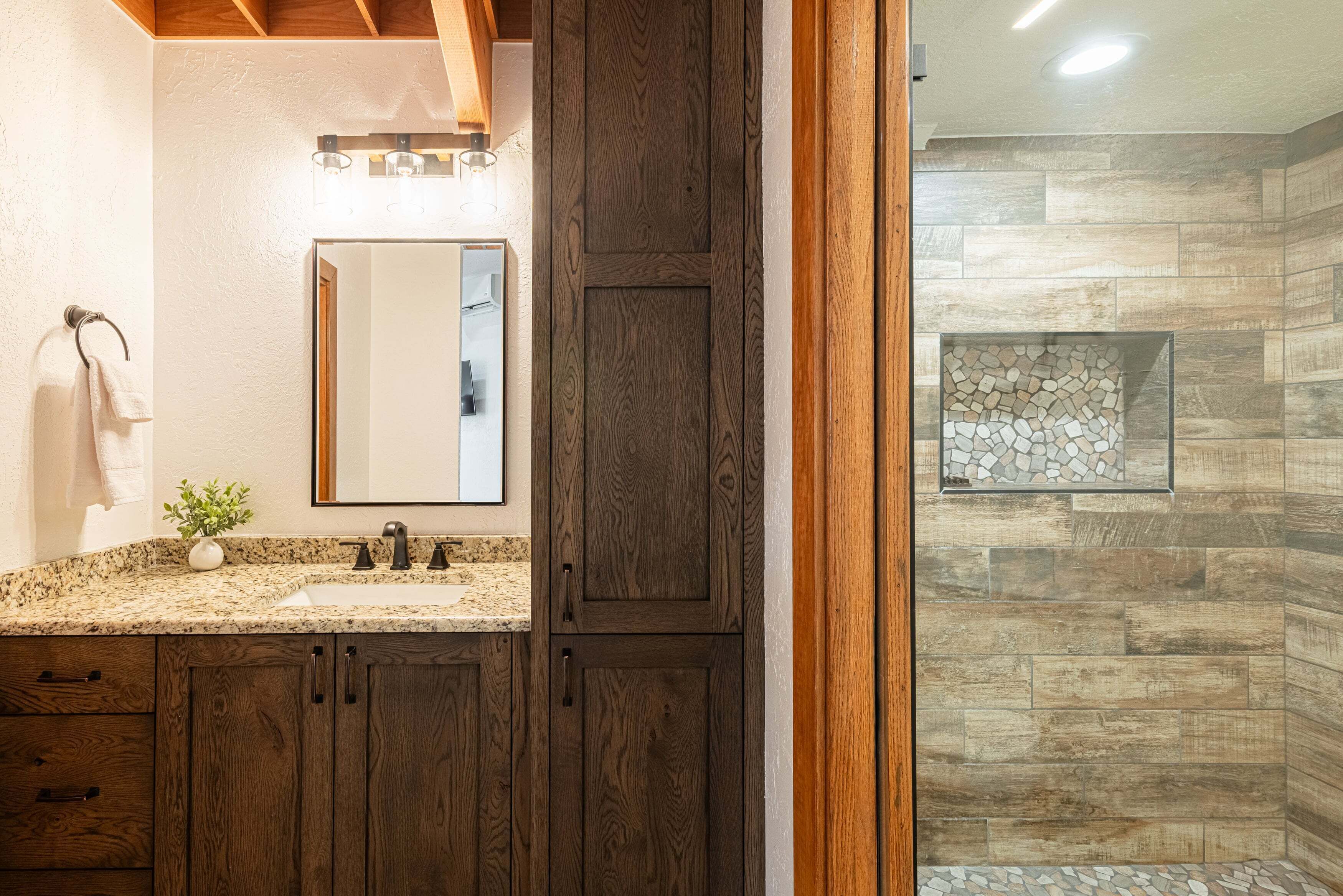 Un baño con encimera de granito, armarios de madera, un espejo, toallero y una ducha con azulejos de piedra.