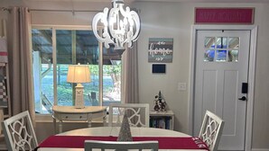 Uma área de refeições com lustre, cadeiras brancas, mesa com toalha vermelha e um letreiro na porta com a mensagem 'Happy Holidays'.