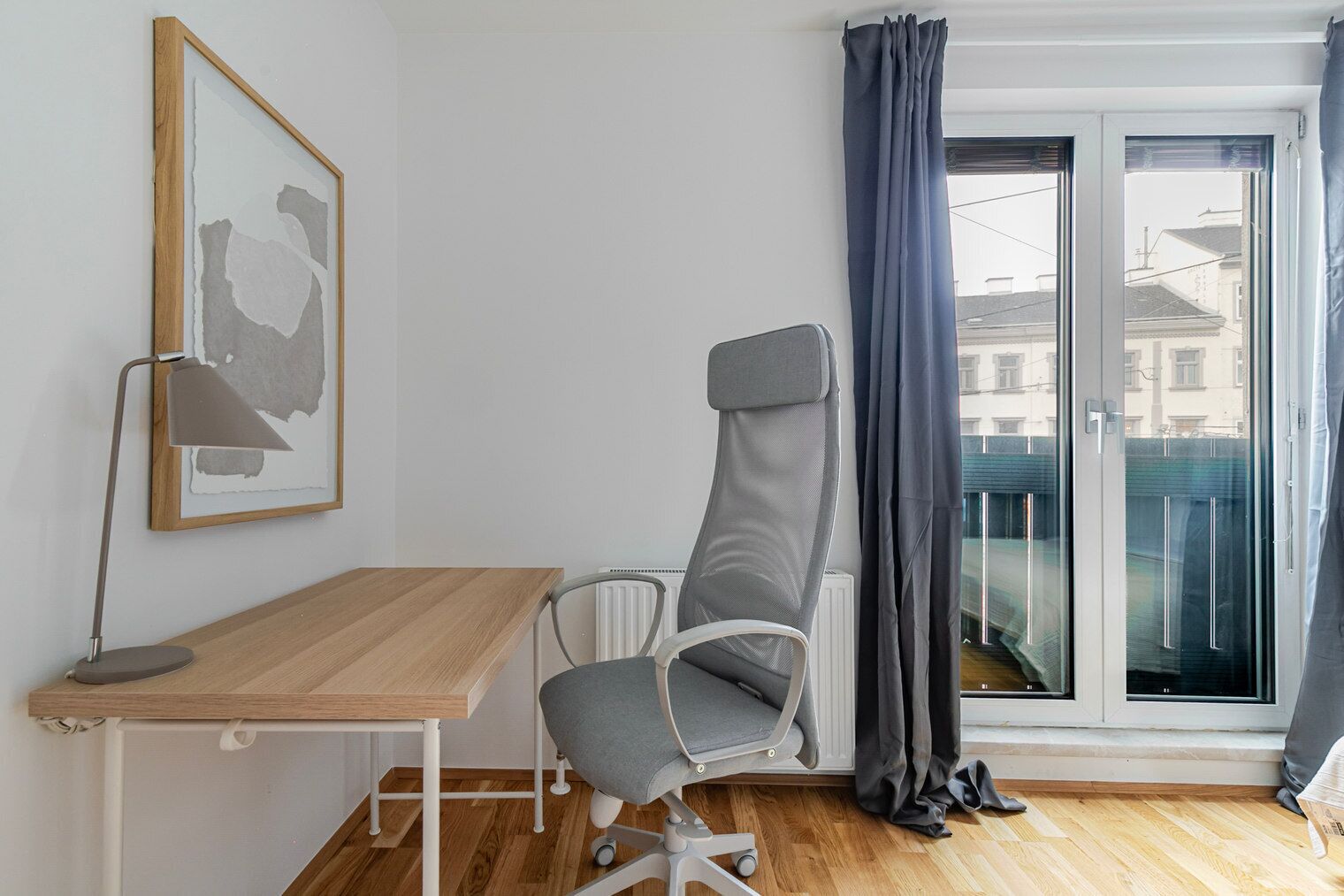 Une pièce avec un bureau en bois, une chaise de bureau grise, un tableau encadré au mur et une vue à travers une porte vitrée.