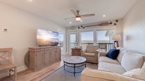 A spacious living room with a large flat-screen TV, a ceiling fan, and a view of the ocean through large windows.