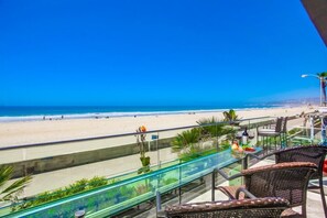 A balcony with wicker chairs, a glass table, and a view of a beach with people and palm trees. - 3 Ocean Front Units * 3 Patios * 2 Levels (San Diego)