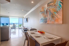 A dining area with a white table set for four, a fruit bowl, and a large tropical leaf painting on the wall. - 3 Ocean Front Units * 3 Patios * 2 Levels (San Diego)