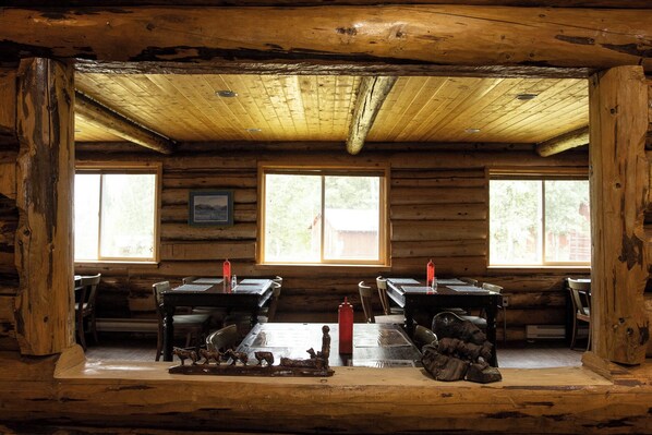 A rustic cabin interior with wooden walls and ceiling, two windows, a dining table with chairs, and a fireplace with logs. - Tatogga Lake Resort (Iskut)