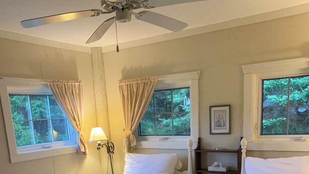 Quarto com duas camas brancas, um ventilador de teto e janelas com cortinas.