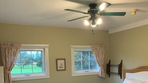 Quarto com cama, duas janelas, ventilador de teto e uma cadeira.