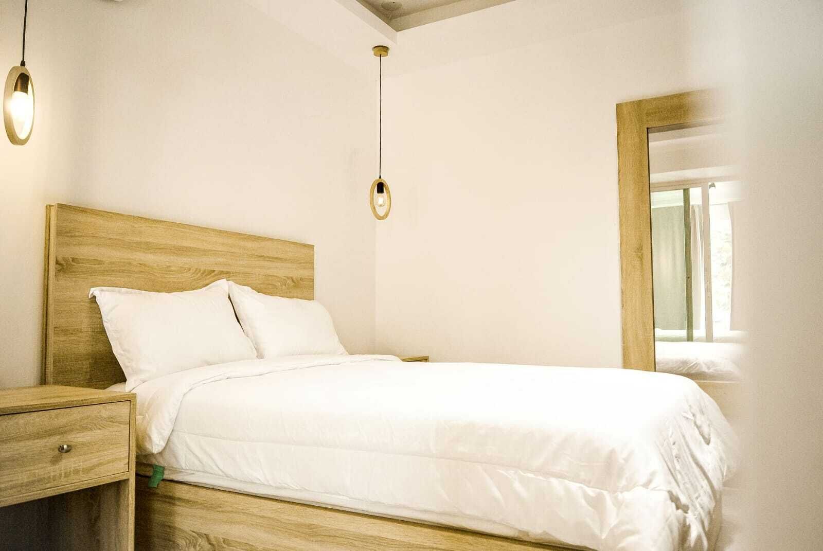 A neatly made bed with a wooden headboard, a bedside table with a drawer, and two pendant lights.