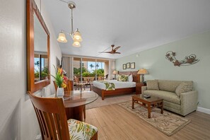 Ein Hotelzimmer mit Bett, einer Couch, einer Essecke mit Tisch und Stühlen, einem Deckenventilator und einem großen Fenster mit Blick auf Palmen.
