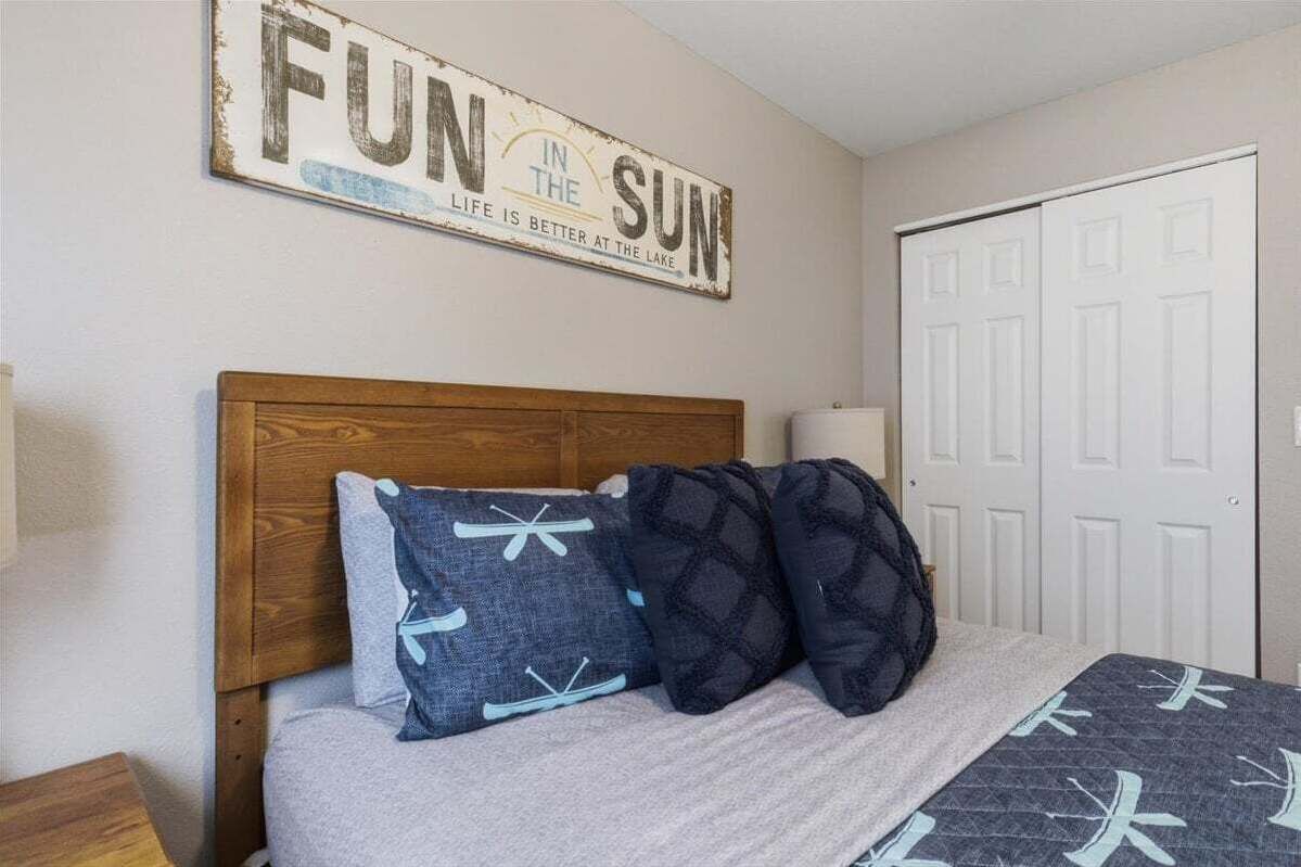 A bedroom with a wooden headboard, a bed with grey and blue patterned bedding, and a wall sign that reads "FUN IN THE SUN".