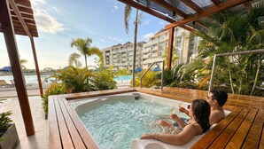 Eine Holzterrasse mit Whirlpool, umgeben von Palmen und Blick auf ein Hotelgebäude.