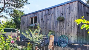Ein rustikales Holzhaus mit Veranda, Schubkarre und einem Garten mit verschiedenen Pflanzen und Blumen.