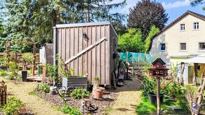 Eine Holzhütte mit geneigtem Dach, umgeben von einem Garten mit verschiedenen Pflanzen und einem kleinen Haus im Hintergrund.