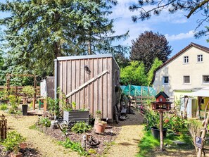 A wooden structure with a slanted roof, surrounded by a garden with various plants and a small house in the background. - Tiny House Cottage Herbert by Interhome (Grossschönau)