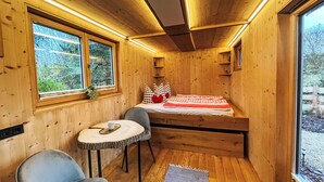 Ein gemütliches Blockhaus-Zimmer mit einem Bett, einem kleinen Tisch, zwei Stühlen und einem Fenster mit Blick auf Bäume.