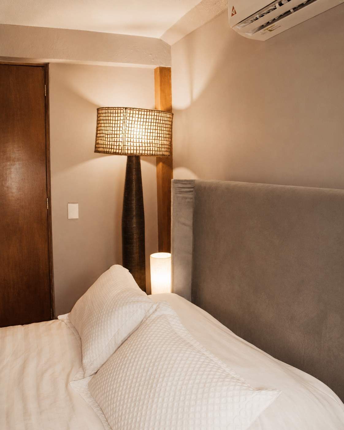 A hotel room with a bed, a nightstand, a lamp, and an air conditioning unit.