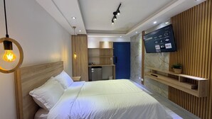 Moderni hotellihuone, jossa on suuri sänky, seinälle asennettu televisio, pieni keittokomero ja sisäänrakennetut puuhyllyt.