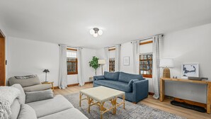 Ein modernes Wohnzimmer mit einem grauen Sofa, einem blauen Sofa, einem Korbtisch und einem Holznachttisch mit einer Lampe. Zwei Fenster mit Vorhängen, eine Pflanze und ein gerahmtes Bild an der Wand.