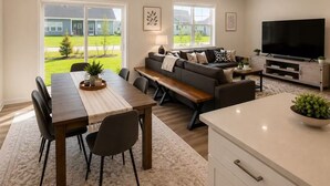A modern living room with a dining area, a sofa, a TV, and large windows. - New 2025 Beach Home! Ocean View Beach Club! 5 Minutes to Bethany Beach (Ocean View)