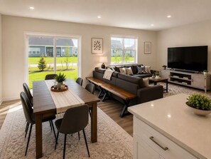 A modern living room with a dining area, a sofa, a TV, and large windows. - New 2025 Beach Home! Ocean View Beach Club! 5 Minutes to Bethany Beach (Ocean View)