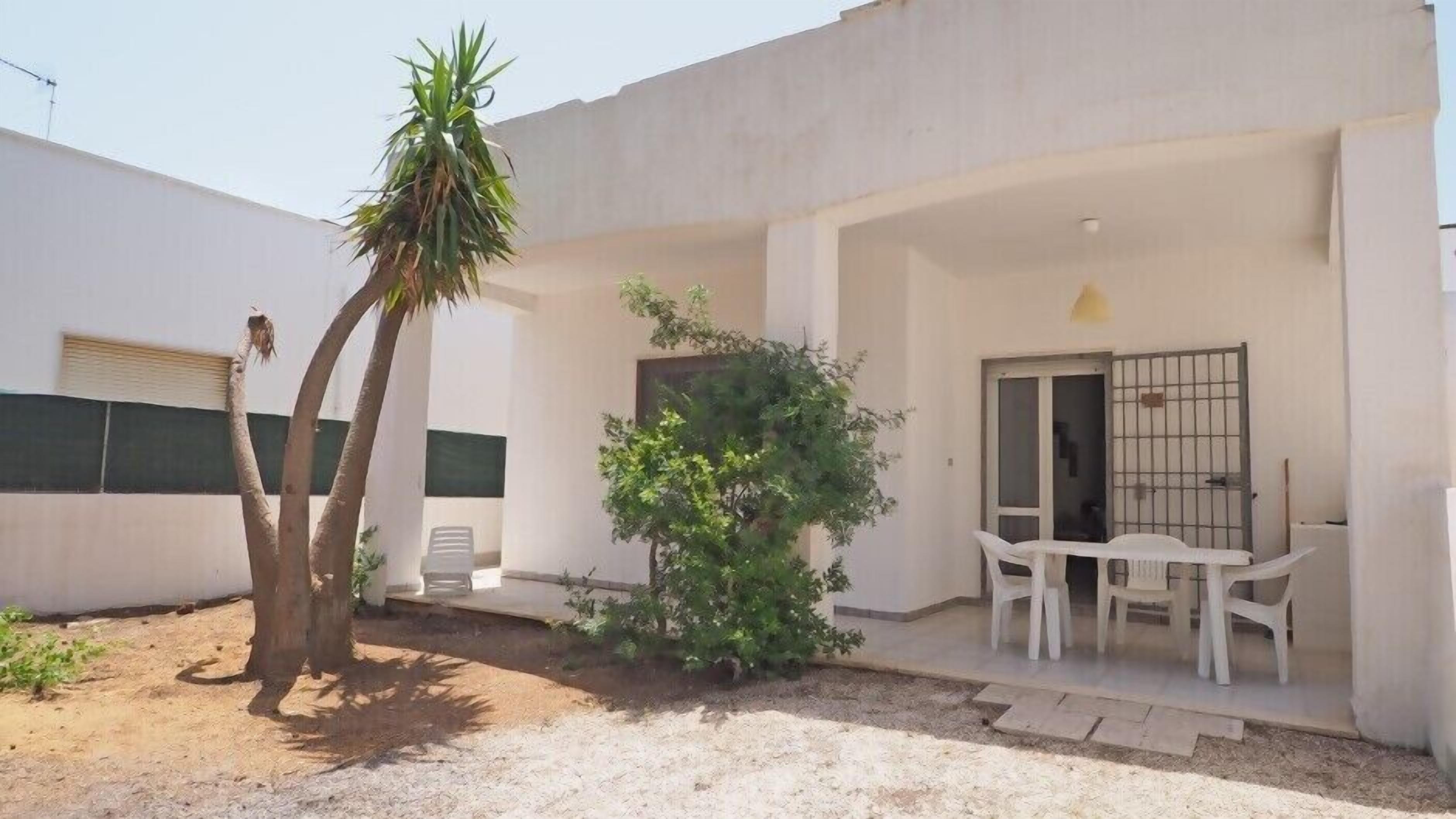 A small, single-story building with a patio area featuring a table and chairs, a small tree, and a door with a metal gate.