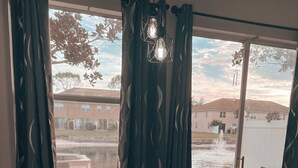 Una habitación con una laptop sobre una mesa, una ventana con cortinas, y vista a un edificio y una masa de agua desde el exterior.