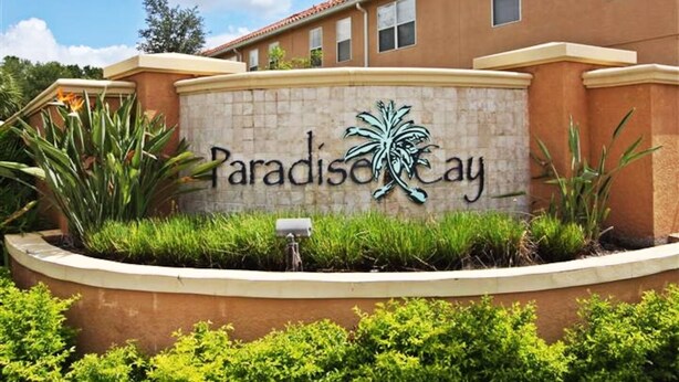 Señal de Paradise Cay con logotipo de palmera, rodeada de exuberante follaje y un paisaje cuidado.