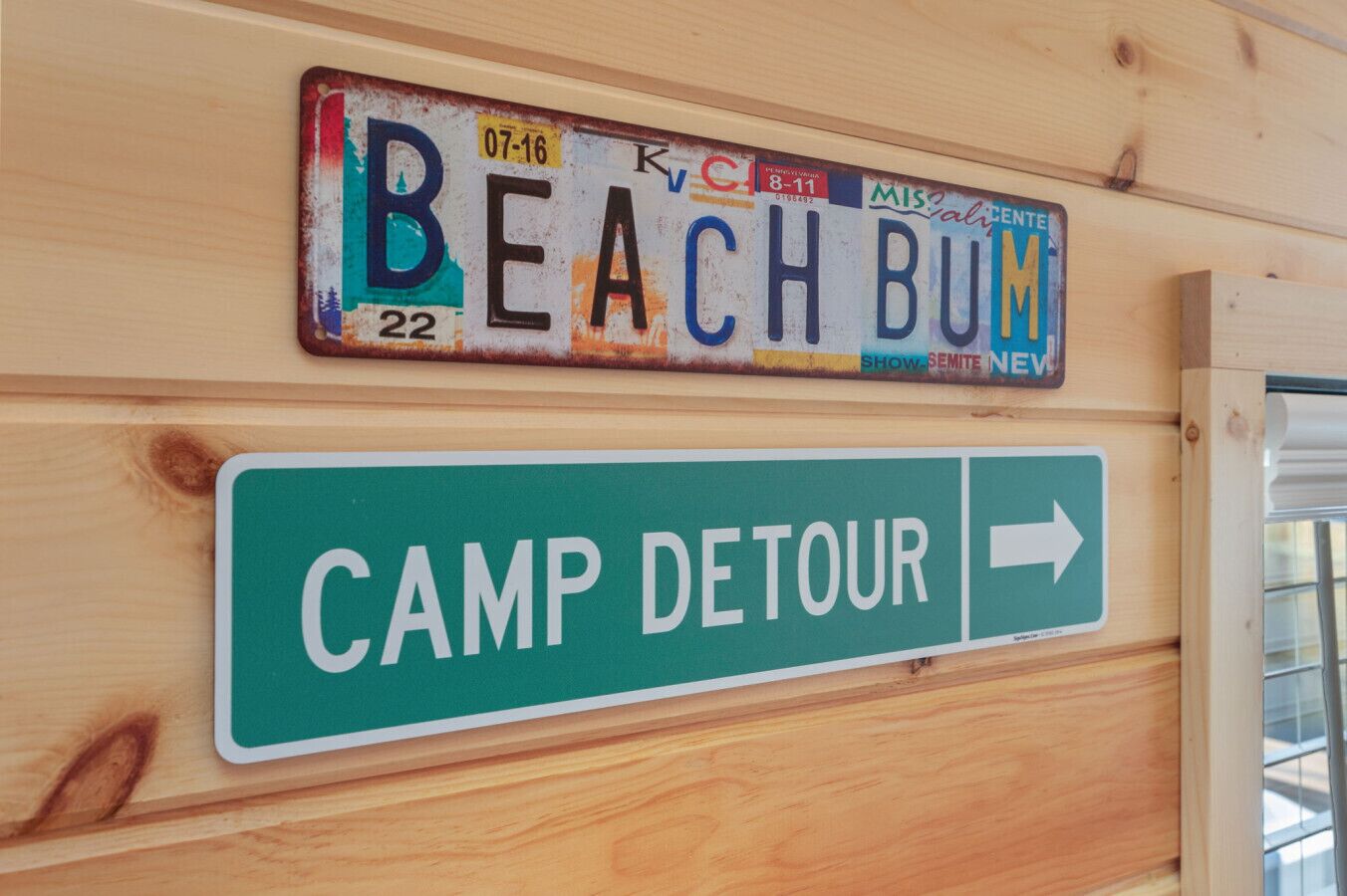 Un mur de cabane en bois orné d’une plaque d’immatriculation sur laquelle on peut lire « BEACH BUM » et d’un panneau indicateur pointant vers « CAMP DETOUR ».