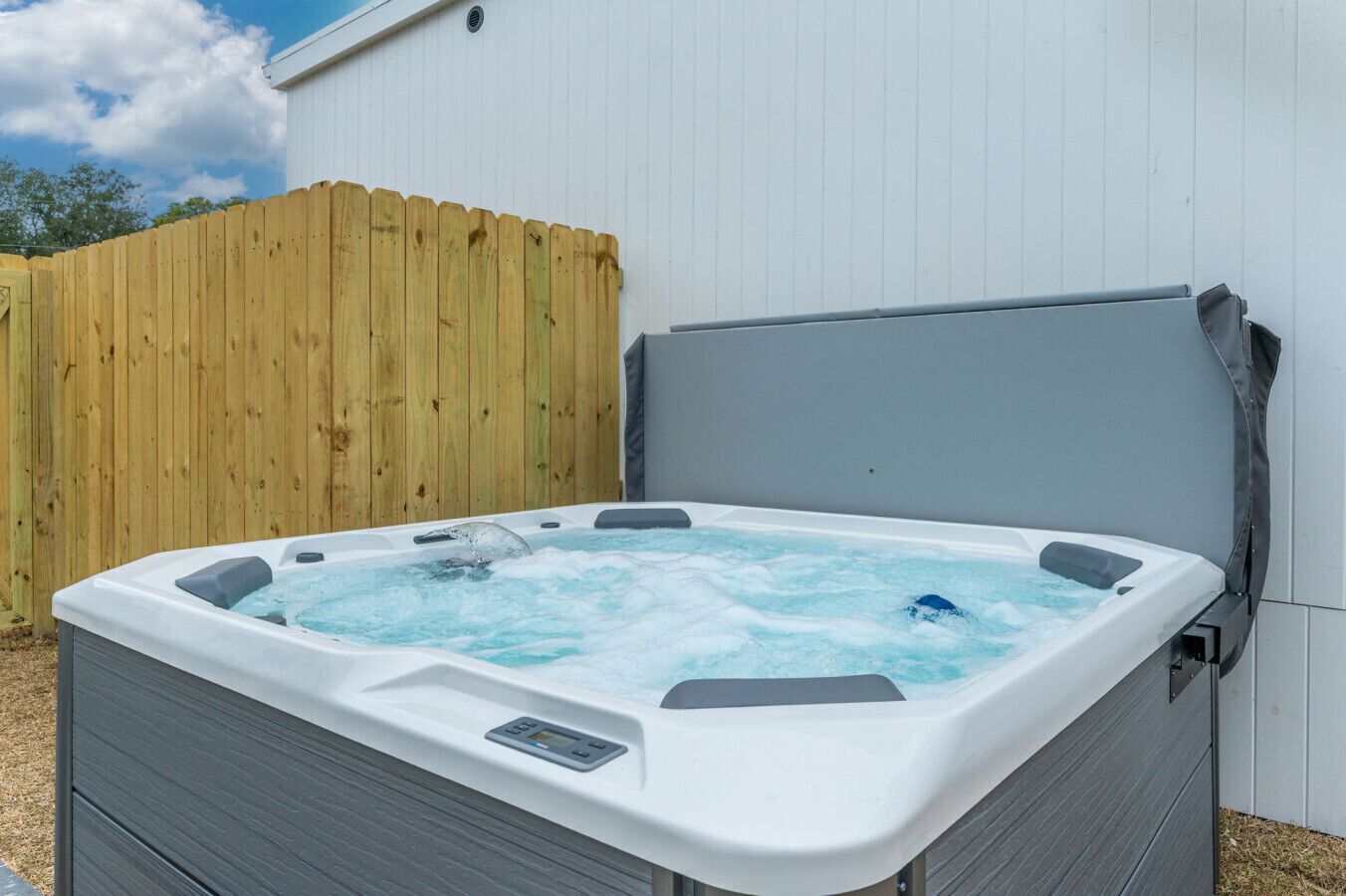 Un bain à remous doté d’un panneau de commande, installé devant une clôture en bois et un bâtiment blanc.