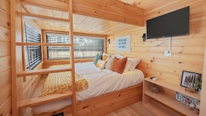 Habitación de cabaña de madera con literas, un televisor montado en la pared y una ventana con persianas.