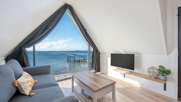 A modern living room with a large window overlooking a body of water, a flat-screen TV, and a wooden coffee table.