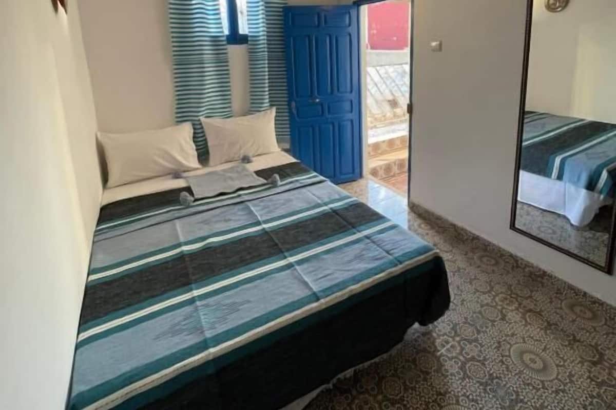 Un dormitori amb un llit, una finestra amb cortines, una porta, una làmpada penjada a la paret i un mirall.