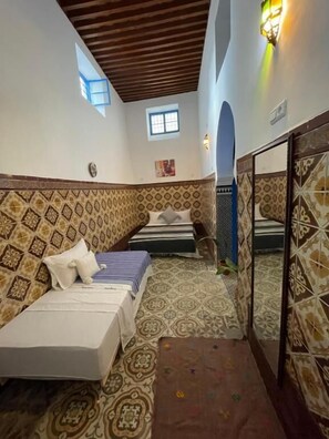 A room with a high ceiling, patterned walls, and a small window. There are two beds with pillows and blankets, a mirror, and a tiled floor.
