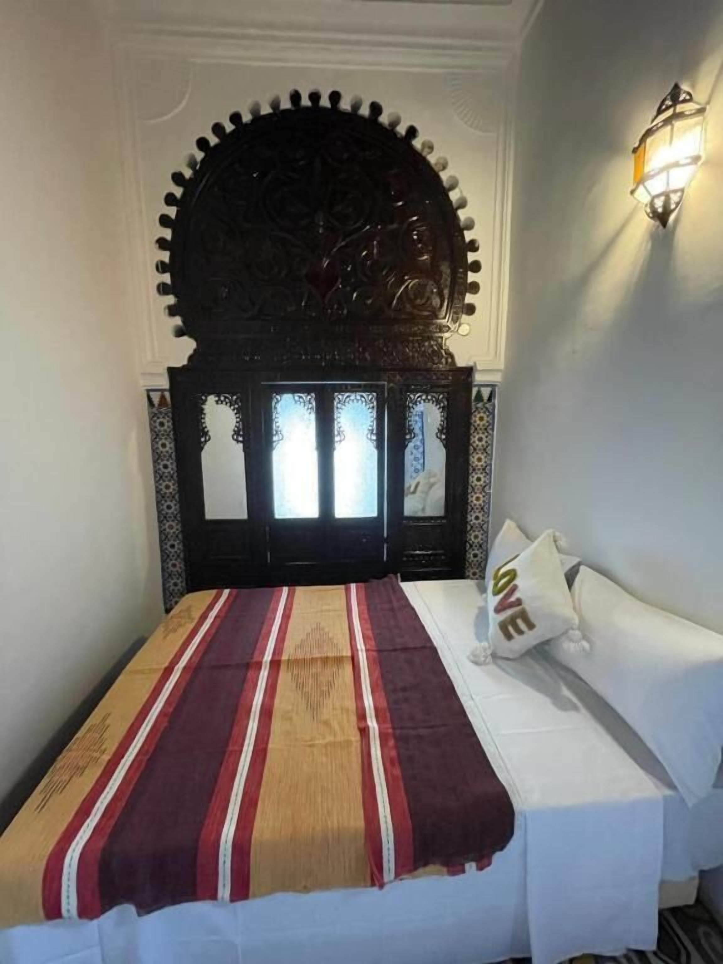 A bedroom with a bed, a decorative headboard, a wall-mounted light fixture, and a patterned bedspread.