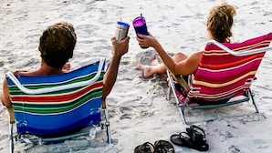 Two individuals seated on beach chairs with striped beach towels, holding drinks, and wearing sandals.
