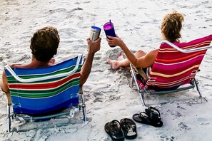 Dos personas sentadas en sillas de playa con toallas a rayas, sosteniendo bebidas y usando sandalias sobre la arena.