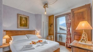 A hotel room with a large bed, a wooden headboard, a painting on the wall, a window with curtains, and a wooden nightstand with a lamp. - Luna Apartment Baita Ayana with Balcony Views, Oulx, Italy (Oulx)
